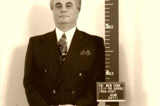 A colour FBI booking photograph of John Gotti in a double-breasted pinstripe suit, standing against a height chart beside a placard reading FBI New York, 12-11-90
