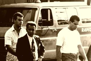 A grainy black and white surveillance photograph of three men walking past a parked van, the older man in the centre wearing a dark suit