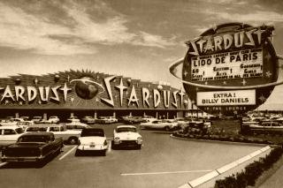A postcard photograph of the Stardust hotel and casino in the early 1960s, its long neon frontage and round roadside sign above a car park full of period cars